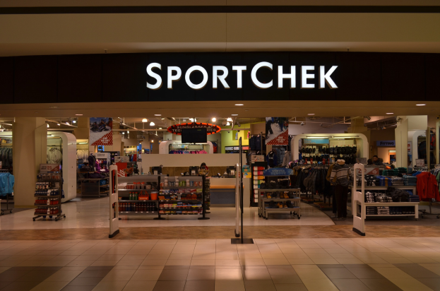 Außen- und Innenansicht eines Sportchek-Geschäfts in einem Einkaufszentrum mit Kleiderständern, Deckenlampen, Pfeilern und einem montierten Fernseher.
