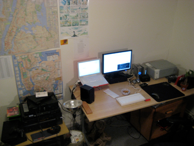 Ein Raum mit einem Tisch, auf dem ein Laptop, ein Lautsprecher, ein Monitor, eine Tastatur, ein Ball, eine Maus, ein Scanner und Papiere liegen, sowie ein weiterer Tisch mit Elektronikgeräten im Hintergrund und Charts und Poster an der Wand dahinter.