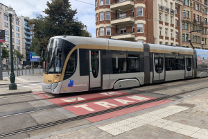 Eine Straßenbahn fährt auf einer von Hochhäusern gesäumten Straße, umgeben von Masten, Laternen, Schildern und Bäumen, mit dem Himmel im Hintergrund.