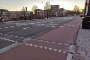 Stadtstraße mit einem ausgewiesenen Fahrradweg, Fahrzeuge, Radfahrer, Laternenpfähle, Bäume, Gebäude mit Fenstern, ein Geländer und sichtbarer Himmel.