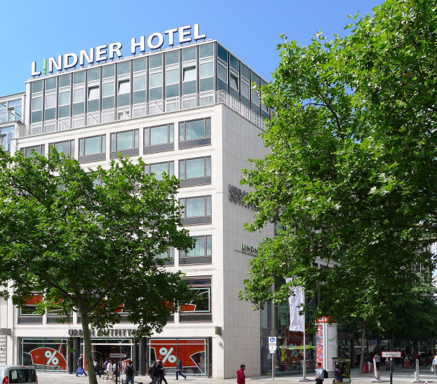 Großes gläsernes Gebäude mit der Aufschrift "Lindner Hotel" in Berlin, umgeben von Bäumen, Fahrzeugen, Passanten und Himmel.
