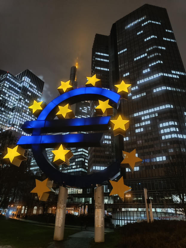 Das Euro-Zeichen steht prominent vor der erleuchteten Zentrale der Europäischen Zentralbank in Frankfurt, Deutschland, umgeben von Bäumen, Pflanzen und Gras, mit dem Himmel im Hintergrund.