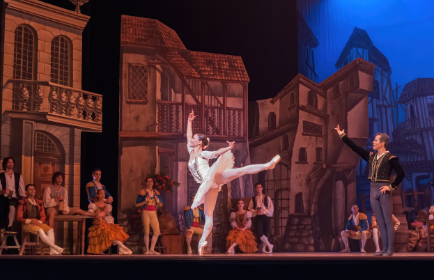 Ein Mann und eine Frau tanzen auf einer Bühne im Royal Opera House in London, mit einer Menschenmenge im Vordergrund und einem Bildschirm mit Gebäudeabbildungen im Hintergrund.