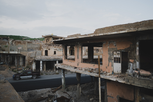 Ein Lastwagen fährt eine Straße neben einem zerstörten Gebäude mit eingestürzten Wänden und verstreuter Trümmer, mit anderen Gebäuden, Pfählen, Bäumen und einem Hügel im Hintergrund.