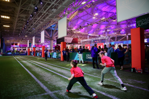 Zwei junge Jungs spielen Fußball auf einem Feld, umgeben von einer Gruppe von Menschen, mit Flaschen und anderen Gegenständen auf der rechten Seite, einem Zaun, Bannern mit Text, einem Anzeigebildschirm, Metallrahmen, einem Dach mit Deckenleuchten und Metallpfosten im Hintergrund.