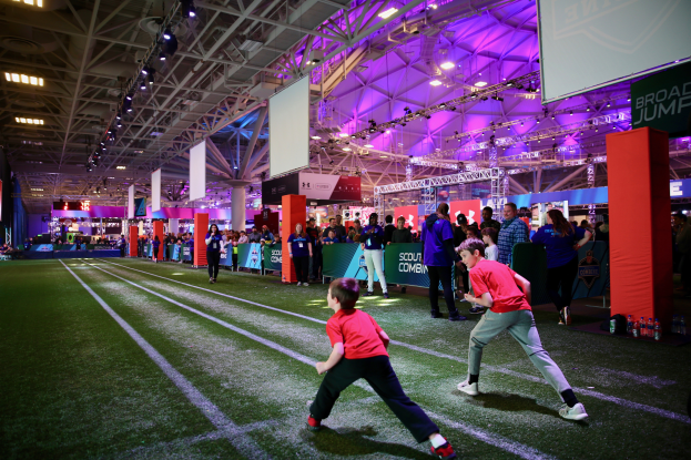 Zwei junge Jungs spielen Fußball auf einem Feld, umgeben von einer Gruppe von Menschen, mit Flaschen und anderen Gegenständen auf der rechten Seite, einem Zaun, Bannern mit Text, einem Anzeigebildschirm, Metallrahmen, einem Dach mit Deckenleuchten und Metallpfosten im Hintergrund.
