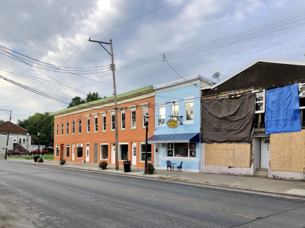 Eine Reihe farbenfroher, zugenagelter Gebäude an einer Straßenecke mit Strommasten, Kabeln, Straßenlaternen, Fahrzeugen, Stühlen, Topfpflanzen, Bäumen und einem bewölkten Himmel.