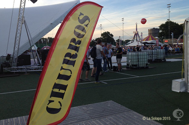 Ein buntes Karnevalszenario mit Zelten, einem zentralen Mast, Menschen, Bäumen, Strommasten, Straßenbahnen auf der Straße und einem eisernen Stand auf der linken Seite unter einem klaren Himmel.