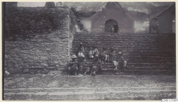 Eine Gruppe von Menschen sitzt auf den Stufen eines Geb├Ąudes, mit einer Wand auf der linken Seite und einer H├╝tte im Hintergrund, und Text am unteren Rand des Bildes.