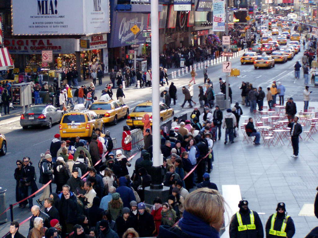 Fahrzeuge passieren eine Straße mit Fußgängern auf beiden Seiten, in der Nähe von Stühlen, Tischen, Laternen, Schildern, Ampeln und Verkehrsschildern, flankiert von Gebäuden mit Geschäften und Namensschildern darüber.
