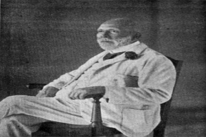 Ein Schwarz-Weiß-Foto eines Mannes in einem Anzug, der in einem Stuhl sitzt und einen ernsten Gesichtsausdruck hat, mit gekreuzten Beinen und Händen auf den Armlehnen, identifiziert als Soziologe im Text unten.