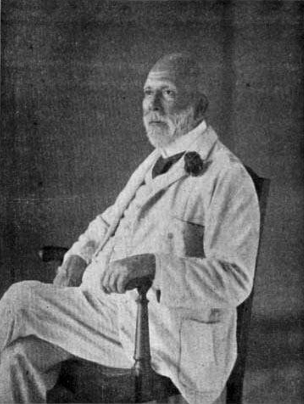 Ein Schwarz-Weiß-Foto eines Mannes in einem Anzug, der in einem Stuhl sitzt und einen ernsten Gesichtsausdruck hat, mit gekreuzten Beinen und Händen auf den Armlehnen, identifiziert als Soziologe im Text unten.