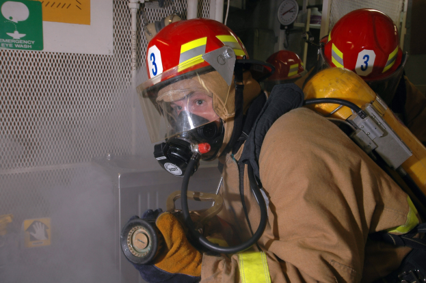 Ein Feuerwehrmann in voller Schutzausrüstung, einschließlich einer Gasmaske, Helm, Handschuhen und Jacke, steht mit Rohren, einer Uhr und anderen Gegenständen im Hintergrund und einer Tafel mit Text auf der linken Seite.