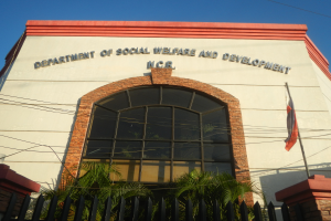 Das Gebäude des Sozialhilfeministeriums mit Text an der Wand, ein Fahnenmast mit einer flatternden Fahne, ein Metallzaun, Pflanzen, Bäume, Drähte und ein bewölkter Himmel.