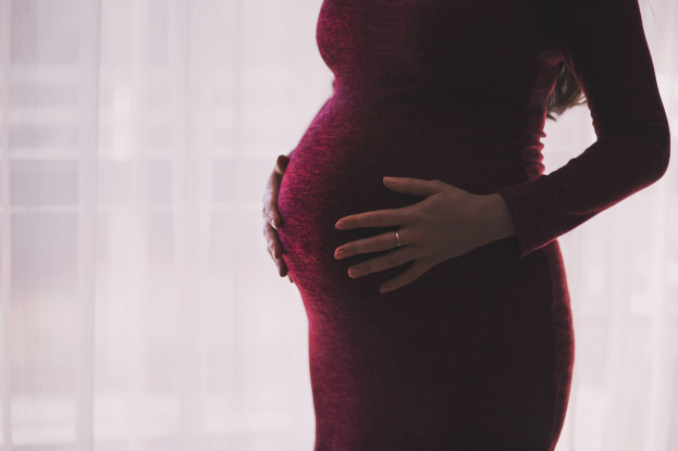 Schwangere Frau in einem roten Kleid, die vor einem Fenster steht, Hände auf dem Bauch, mit einem leicht unscharfen Hintergrund.