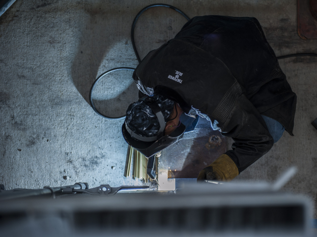 Ein Schweißer in Schutzausrüstung bedient eine Schweißmaschine an einem Metallstück, umgeben von Werkzeugen und Gegenständen auf dem Boden.