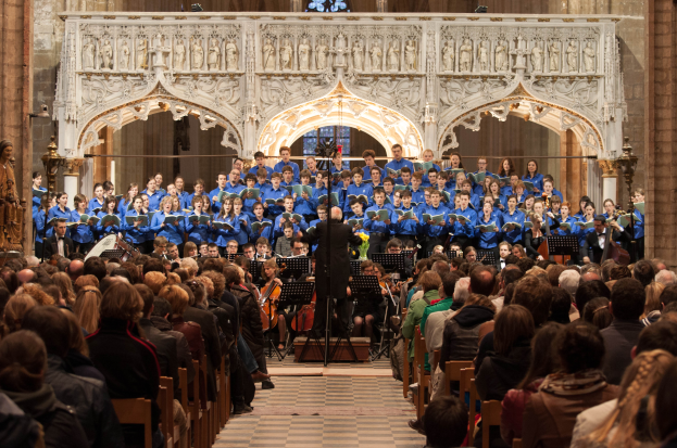 Eine große Gruppe von Menschen steht vor einem Chor in einer Kirche, mit einer Person in der Mitte, die ein Mikrofon hält, sowie Skulpturen und Statuen im Hintergrund.