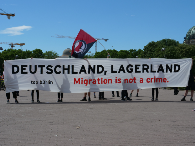 Eine Gruppe von Menschen mit einem Banner, auf dem "Deutschland, Lagerland Migration ist kein Verbrechen" steht, und einer Flagge, die auf dem Boden steht, mit Straßenlaternen, Bäumen, Kränen, einem Gebäude und einem bewölkten Himmel im Hintergrund.