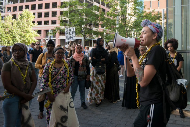 Eine Frau in einem Kopftuch, T-Shirt, Hose und einer Tasche hält einen Lautsprecher und steht zwischen vielen Frauen in Burkas, umgeben von Bäumen, Polen, Gebäuden und einer Gruppe von Menschen in der Nähe eines Plakats.