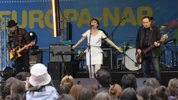 Eine Bühne mit vier Musikern: eine singende Frau in weißem Kleid in der Mitte, zwei Gitarristen links und rechts und eine Menge und Lautsprecher im Hintergrund.