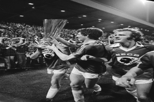 Schwarzes Bild von Fußballspielern, die mit einem Pokal auf einem Feld feiern, umgeben von einer Menge hinter einem Zaun unter Stadionbeleuchtung.