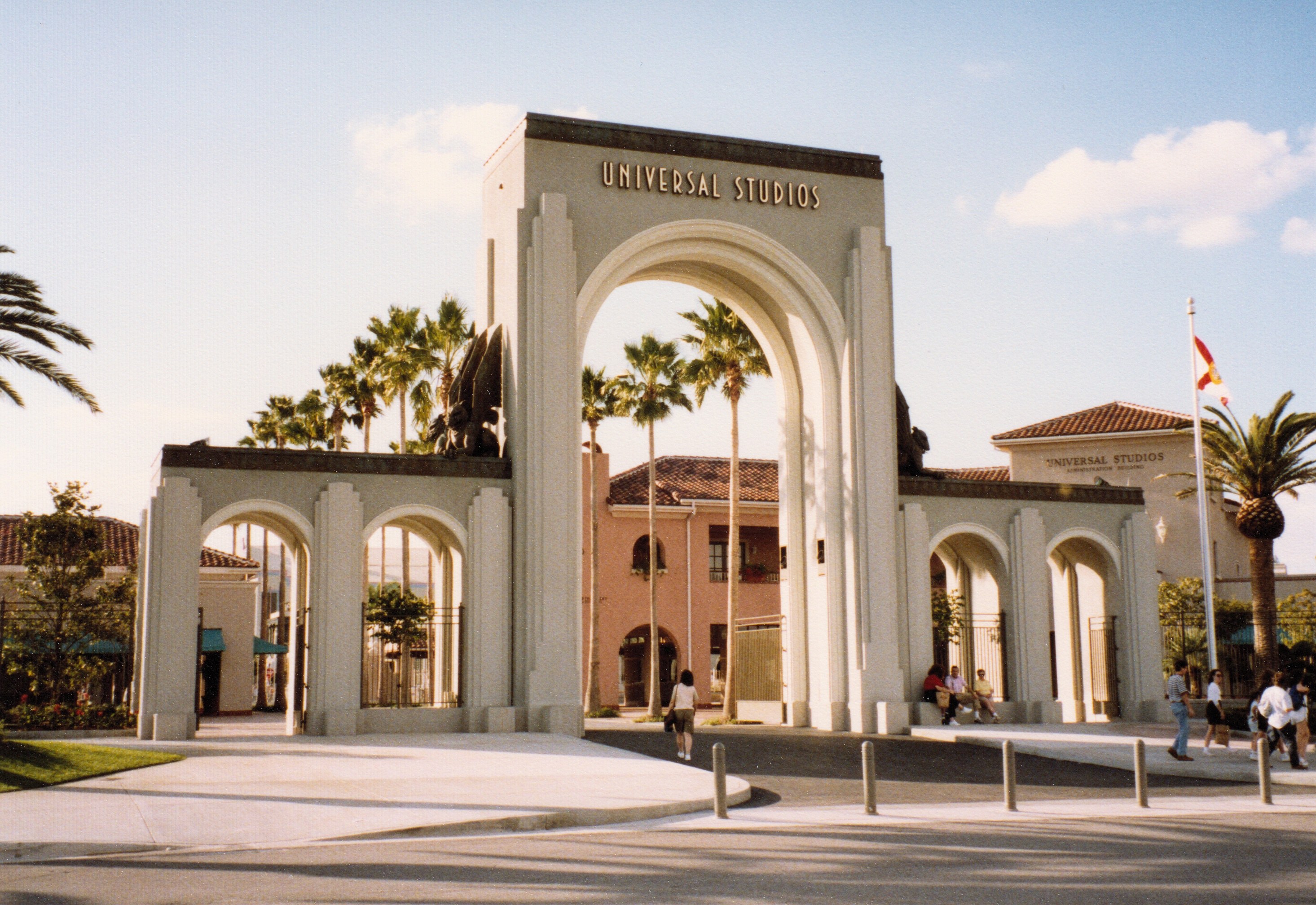 Eingang zu Universal Studios Hollywood mit einem Bogen mit Text, einer Statue, Menschen, umgeben von Grün, Gebäuden, einem Fahnenmast mit einer Fahne.