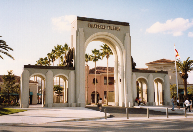 Eingang zu Universal Studios Hollywood mit einem Bogen mit Text, einer Statue, Menschen, umgeben von Grün, Gebäuden, einem Fahnenmast mit einer Fahne.