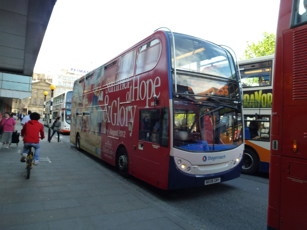 Eine Person fährt mit einem Fahrrad auf einem Gehweg mit vielen Menschen, Bussen mit Text darauf, Gebäuden und Laternen im Hintergrund.