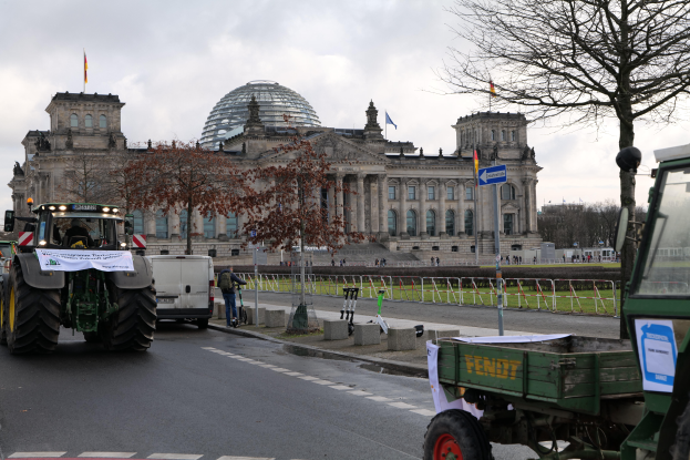 Eine Gruppe von Traktoren fährt vor dem Reichstaggebäude in Berlin, Deutschland, die mit Fenstern, Säulen und Flaggen geschmückt ist und von Bäumen, Geländern, Pfählen und Brettern umgeben ist, mit ein paar Menschen im Hintergrund und einem bewölkten Himmel.