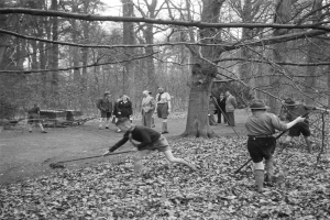 Schwarz-weiß-Foto von Menschen im Freien in einem bewaldeten Gebiet, die mit Stöcken interagieren.