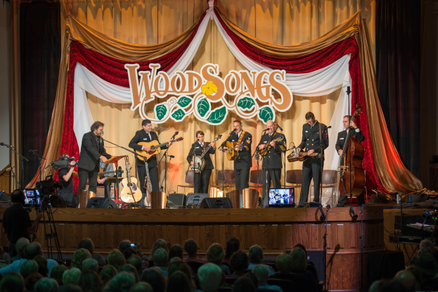Eine Gruppe von Musikern, die auf der Bühne des Wood Songs Festivals spielen, mit Zuschauern vorne und einem Kameramann links.