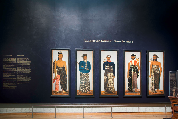 Ein Raum mit einem Holzboden, ein Tisch mit verschiedenen Gegenständen, eine Wand mit Foto Rahmen und dem Text "Javanische Kunst im Nationalmuseum der Niederlande" und eine Vitrine voller Bilder.