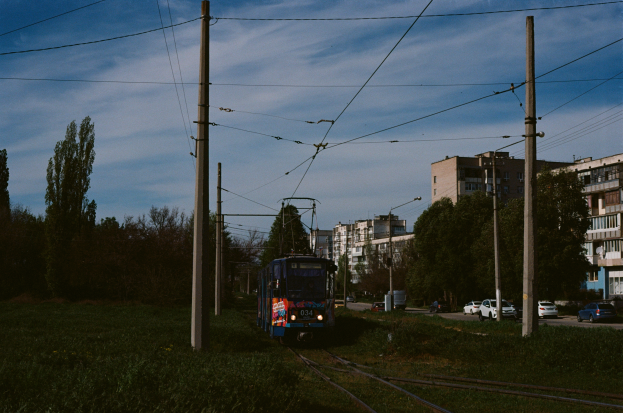 Eine blaue Straßenbahn fährt auf Schienen neben hohen Gebäuden, mit Strommasten, Drähten, Gras und Fahrzeugen auf der Straße; Bäume und Gebäude mit Fenstern sind im Hintergrund unter einem bewölkten Himmel.