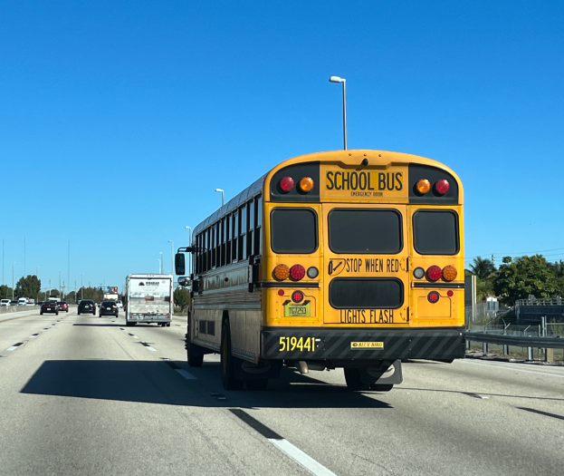 Ein gelber Schulbus fährt auf einer Autobahn, umgeben von anderen Fahrzeugen, mit einer Begrenzung auf der rechten Seite, Bäumen, Pfählen und einem klaren blauen Himmel im Hintergrund.