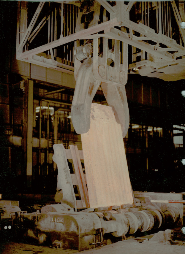 Altes Foto eines großen Industriekrans, der ein Metallförderband in einer Fabrik hochhebt, mit einem Gebäude und einem Metallobjekt im Hintergrund.