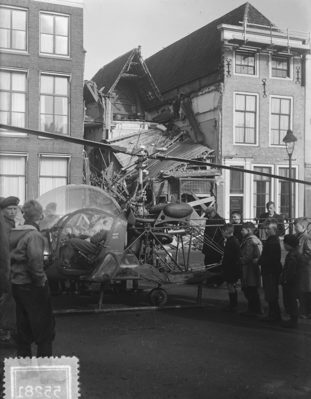 Schwarz-weißes Bild eines kleinen Flugzeugs, das in ein Gebäude gekracht ist, mit Menschen drumherum und einer Person im Fahrzeug, Lichtmast und Fenster am Gebäude, Himmel im Hintergrund.