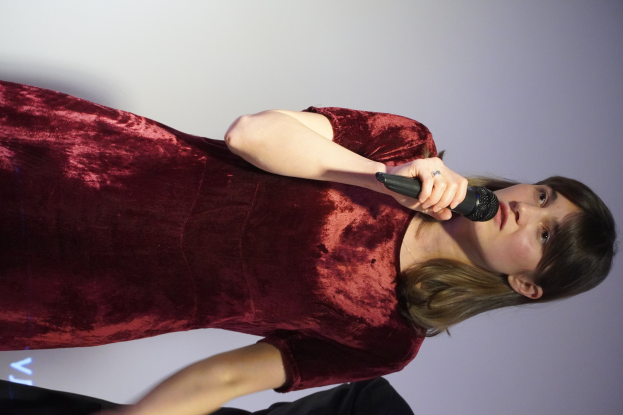 Eine Frau in einem roten Kleid steht selbstbewusst auf einer Bühne, hält ein Mikrofon in der Hand und hat eine sleek Bob-Frisur sowie dezentes, natürliches Make-up.