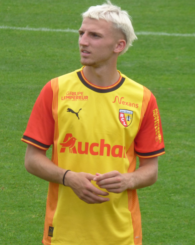 Ein Fußballspieler in einem gelb-orangen gestreiften T-Shirt steht selbstbewusst auf einem Feld und schaut entschlossen nach vorne.