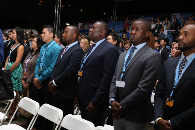 Männer in Blazern und Ausweisen stehen auf einem Boden mit Stühlen vor dunklem Hintergrund mit Absperrgittern und Eisenpfeilern.