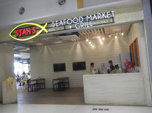 Inneneinrichtung von Stans Seafood Market & Grill mit sitzenden und stehenden Menschen, einem Tisch mit Gegenständen auf der rechten Seite, einem Foto auf der Wand und an der Wand montierten Fernsehern und Lampen.