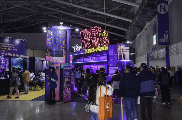 Eine große Gruppe von Menschen steht vor einem Messestand, der als 'Indie Game Development Award 2019' gekennzeichnet ist, wobei einige Taschen und Messestände mit Text, Bildschirmen und Beleuchtung im Hintergrund halten.