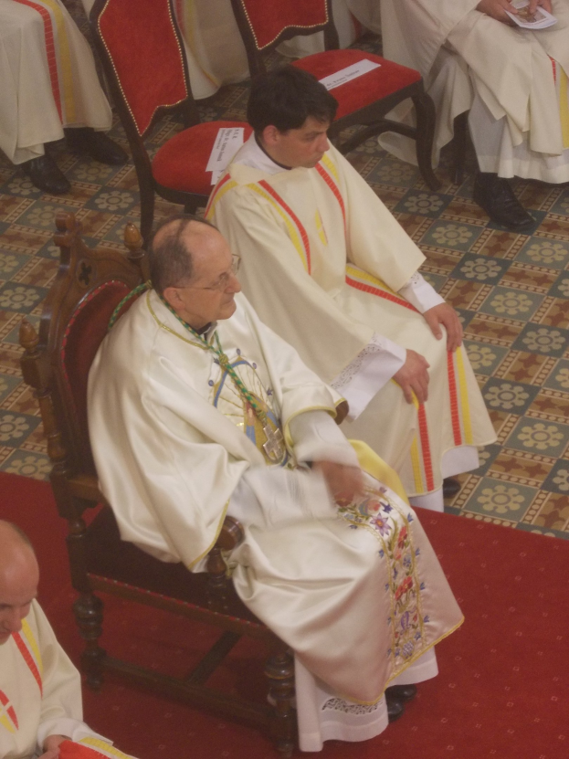 Gruppe von Priestern in weißen Gewändern, die auf Stühlen in einer Kirche sitzen, einige halten Papiere, auf einem roten Teppich.