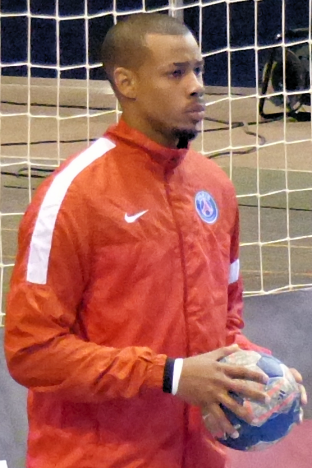 Ein Mann in einem blauen Hemd und Shorts steht vor einem Tor und hält einen Fußball mit entschlossener Miene.