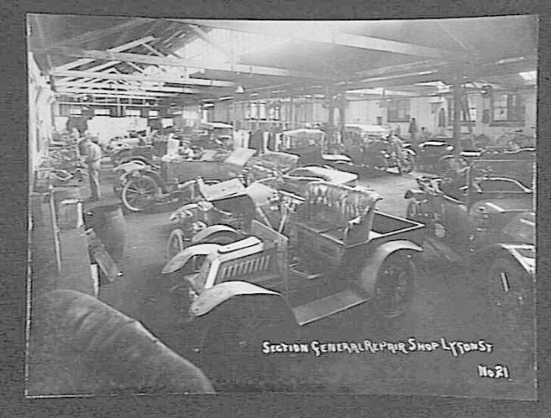 Schwarz-weiß-Foto einer frühen 20. Jahrhundert-Autowerkstatt in Lyon, Frankreich, mit mehreren Fahrzeugen, Menschen und verstreuten Werkzeugen unter einer sichtbaren Decke und Text unten.