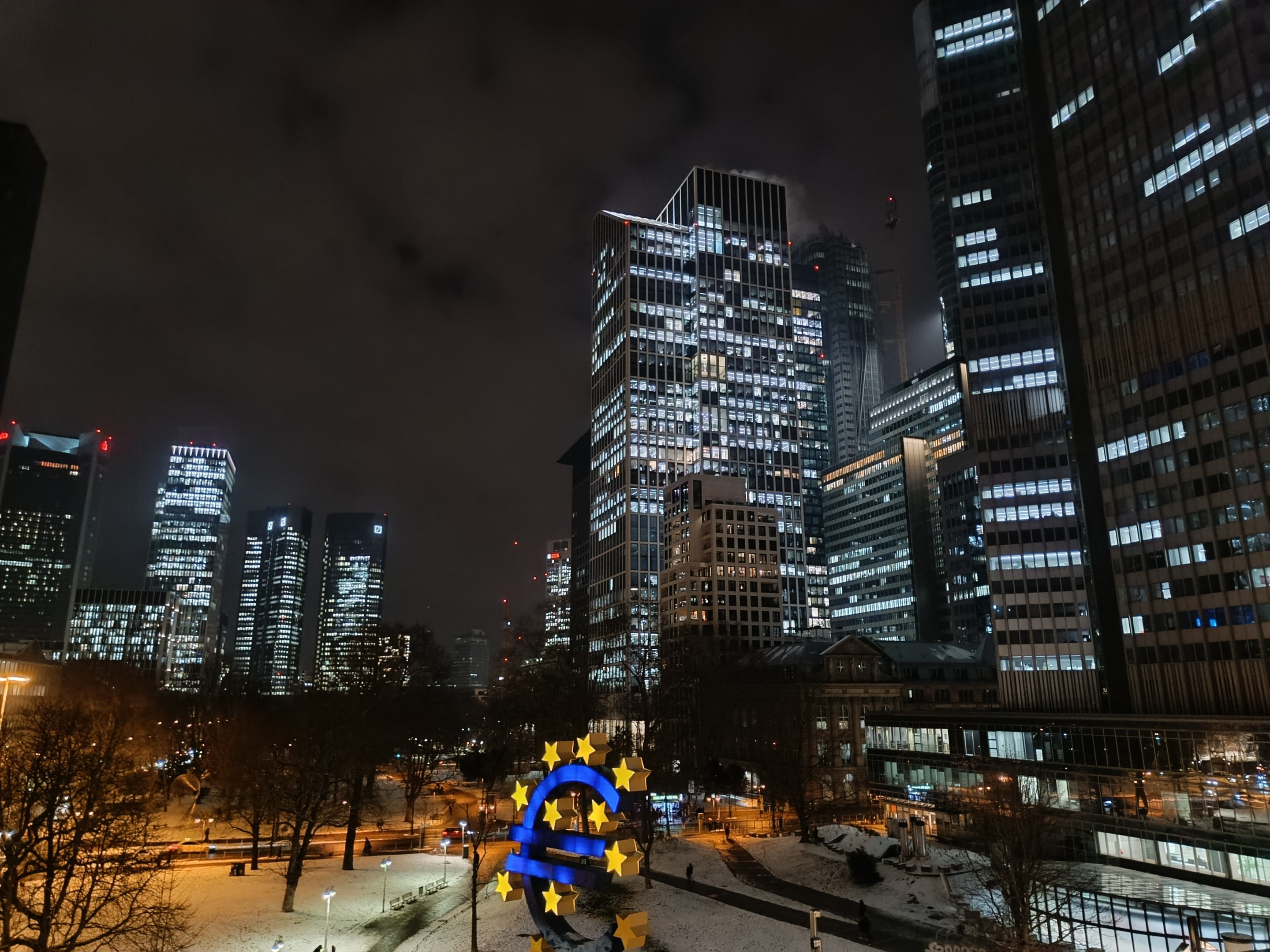 Europäische Zentralbank-Hauptquartier in Frankfurt, Deutschland bei Nacht mit beleuchteten Gebäuden, Bäumen, Straßenlaternen, Lichtern und Menschen, die auf der Straße unter einem bewölkten Himmel gehen.
