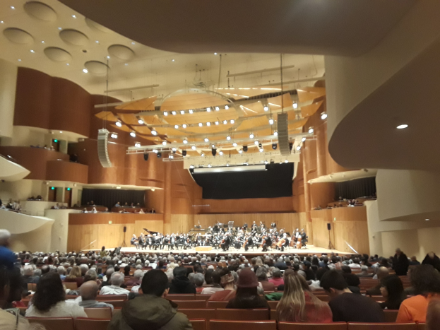 Ein großer Saal voller Menschen, die ein Konzert verfolgen, mit einer Gruppe auf der Bühne, die Instrumente spielt, und anderen, die sitzen, unter einer mit Lichtern geschmückten Decke.