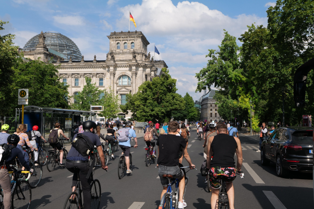 Gruppe von Menschen, die Fahrräder auf einer von Bäumen gesäumten Straße in Berlin, Deutschland, fahren, mit einer Bushaltestelle auf der rechten Seite und einer Flagge auf einem Gebäude unter einem bewölkten Himmel.