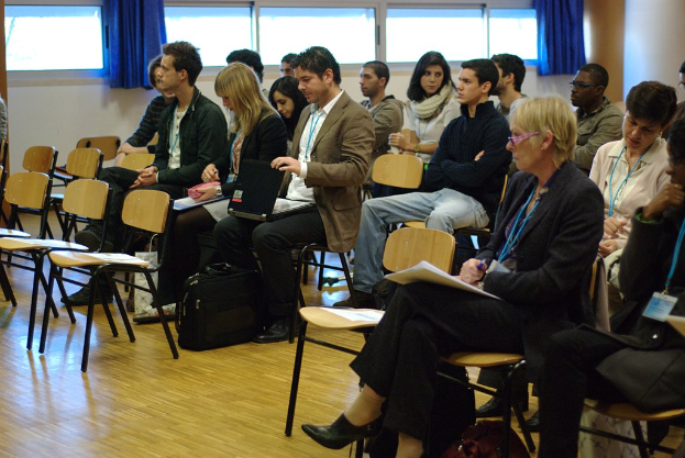 Mehrere Personen auf Stühlen sitzend, einige mit Laptops oder Stiften, mit leeren Stühlen, einer Tasche und Fenstern mit Vorhängen im Hintergrund.