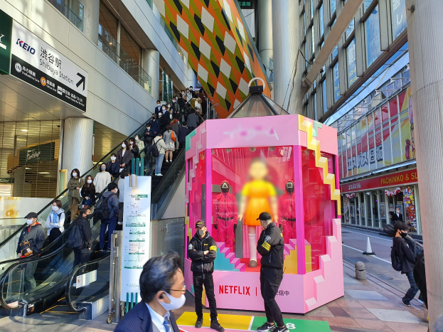 Eine Gruppe von Menschen steht vor einem Gebäude mit Glaswänden und Texttafeln, einige tragen Masken, während eine Rolltreppe mit Menschen zu sehen ist; eine pinkfarbene Box in der Mitte zeigt Schaufensterpuppen und trägt den Text "Netflix's new Netflix booth in Tokyo."