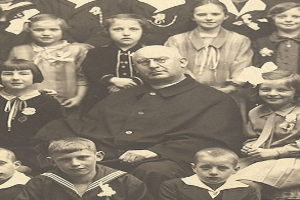 Ein Schwarz-Wei├č-Foto eines strengen Mannes, der in der Mitte sitzt und von einer Gruppe Kinder umgeben ist, die ihn bewundernd anschauen.